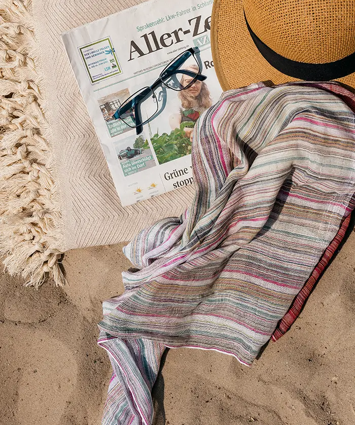 Strandtuch mit bunten Streifen, Strohhut, Sonnenbrille und Zeitung auf Sand am Seeufer
