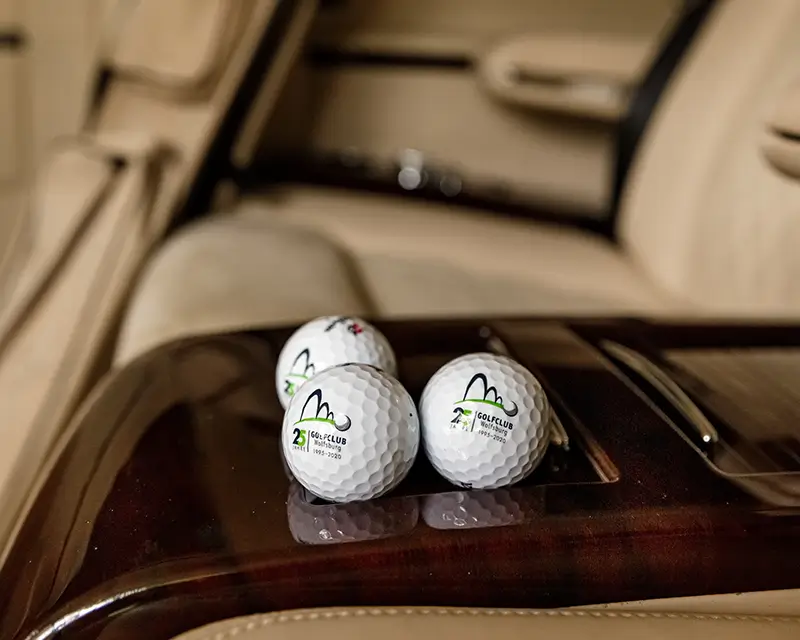 Drei Golfbälle mit Logo auf einer glänzenden Holzarmlehne im Fond eines Luxusautos.