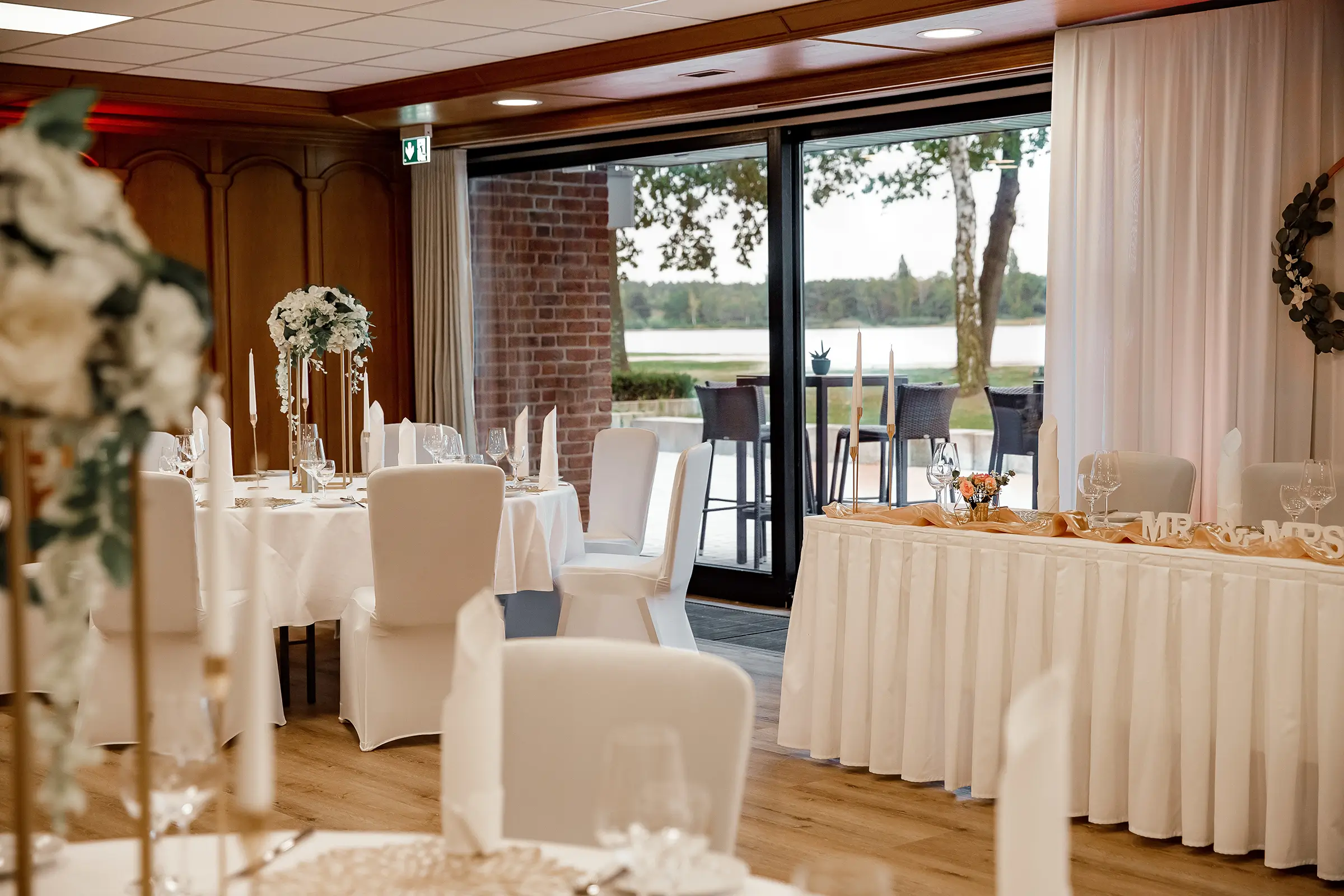 Festlich gedeckte Tische in einem Raum mit Blick auf den Tankumsee, dekoriert für eine Hochzeit mit weißen Stuhlhussen und Blumenschmuck