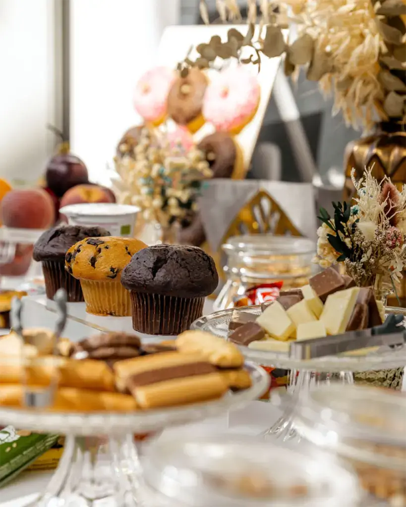 Vielfältiges Buffet mit Muffins, Schokolade, Keksen, Obst und Donuts, dekoriert mit Trockenblumen.