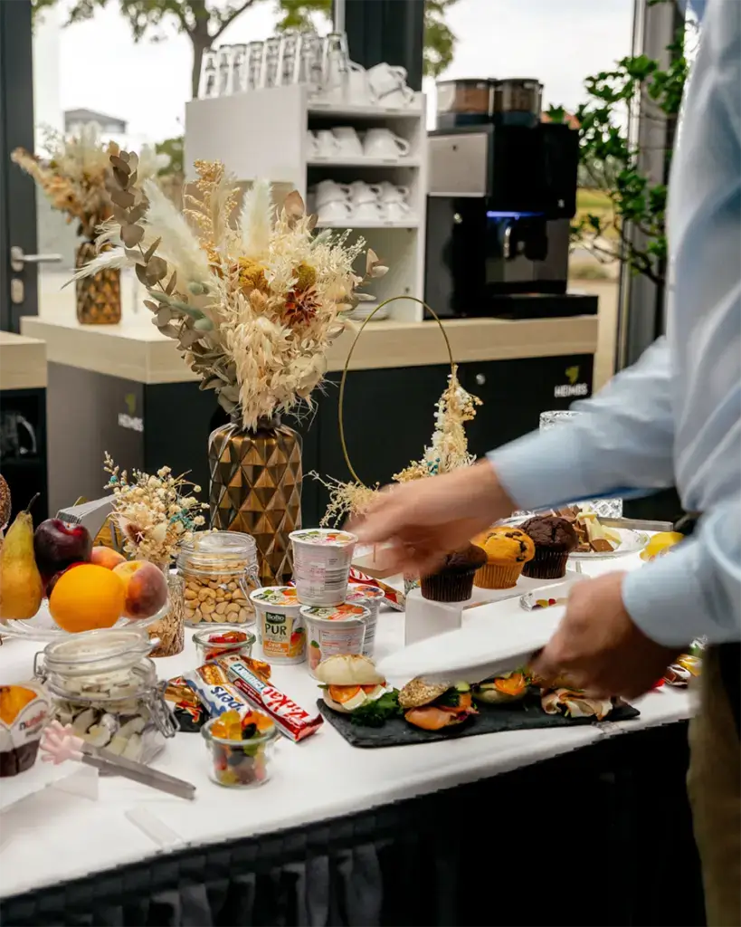 Kaffeepausen-Buffet mit Snacks, belegten Brötchen, Joghurt und frischem Obst, im Hintergrund eine Kaffeemaschine und Trockenblumen-Deko.