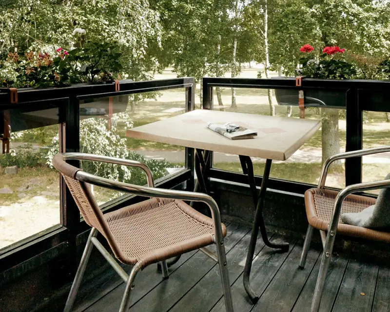 Balkon mit zwei Stühlen aus Metall und geflochtenem Material, quadratischem Tisch mit Buch, umgeben von Glasgeländer und Pflanzen
