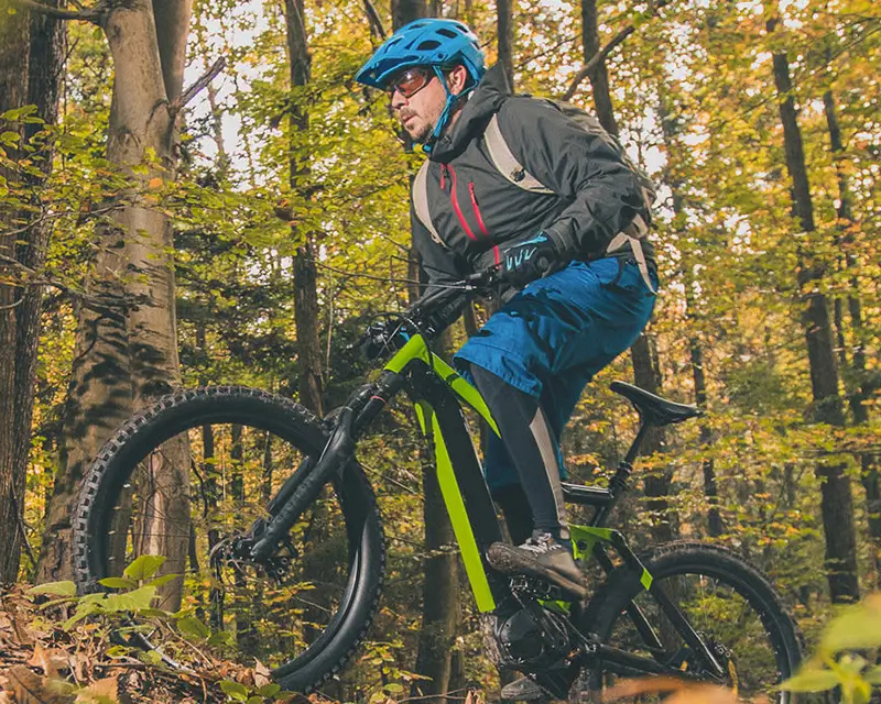 Mountainbiker mit blauem Helm und schwarzer Jacke fährt im Wald auf einem schwarzen und grünen Fahrrad