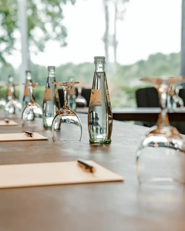 Detailaufnahme eines Konferenztischs mit Wasserflaschen, Gläsern, Blöcken und Stiften, im Hintergrund große Fenster mit Blick ins Grüne.