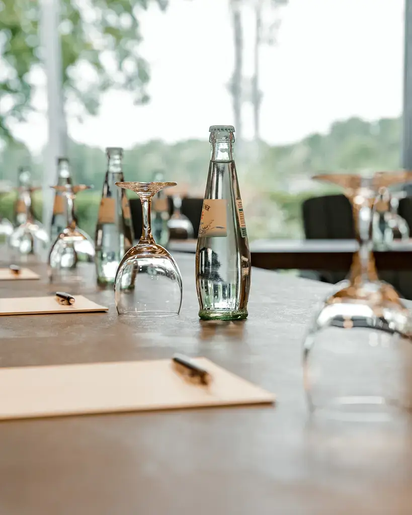 Detailaufnahme eines Konferenztischs mit Wasserflaschen, Gläsern, Blöcken und Stiften, im Hintergrund große Fenster mit Blick ins Grüne.