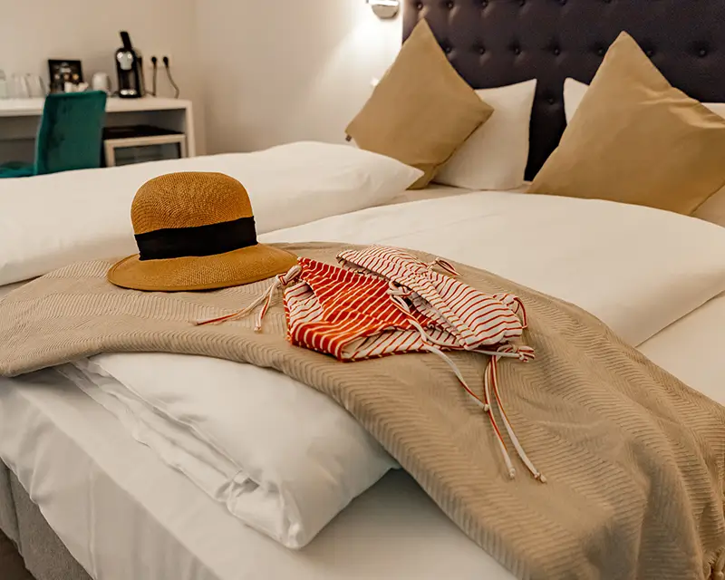 Gemütliches Hotelzimmer mit Doppelbett, darauf ein Strohhut und gestreifte Bikinis – bereit für den Tag am See.